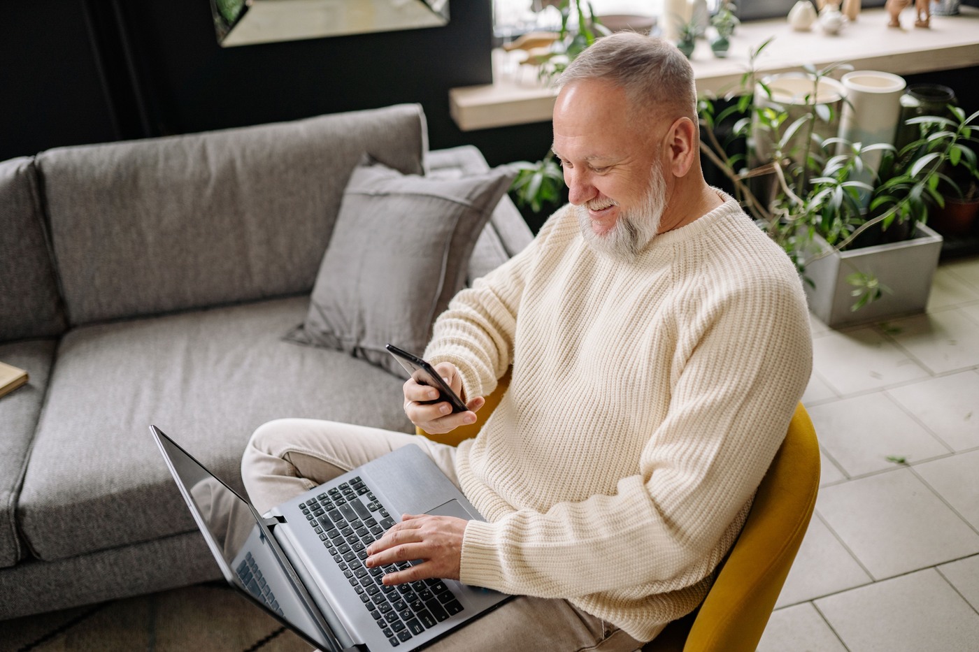 An older man checks his smartphone while sitting at home with a laptop.