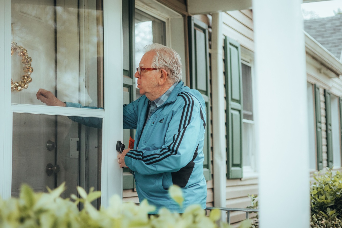 An older man in a blue jacket stands at a house entrance and knocks on the door.