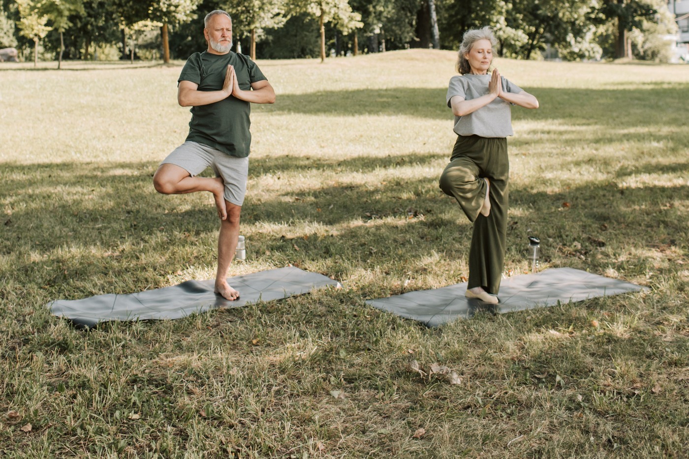 An older woman and man practice standing yoga poses together on mats in a sunny park.